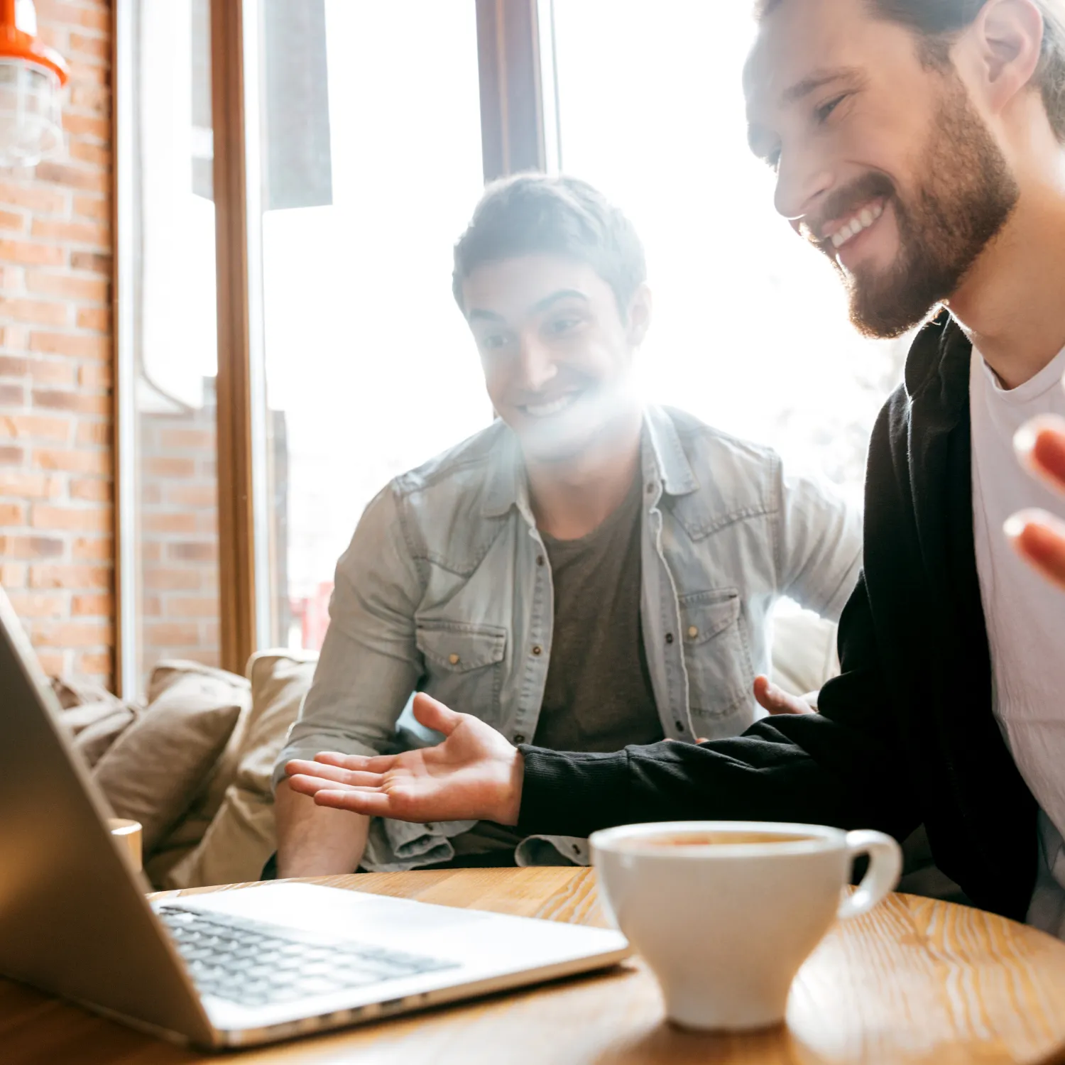 Zwei junge Männer sitzen lächelnd vor einem Laptop in einem Café und unterhalten sich. Eine Kaffeetasse steht auf dem Tisch.