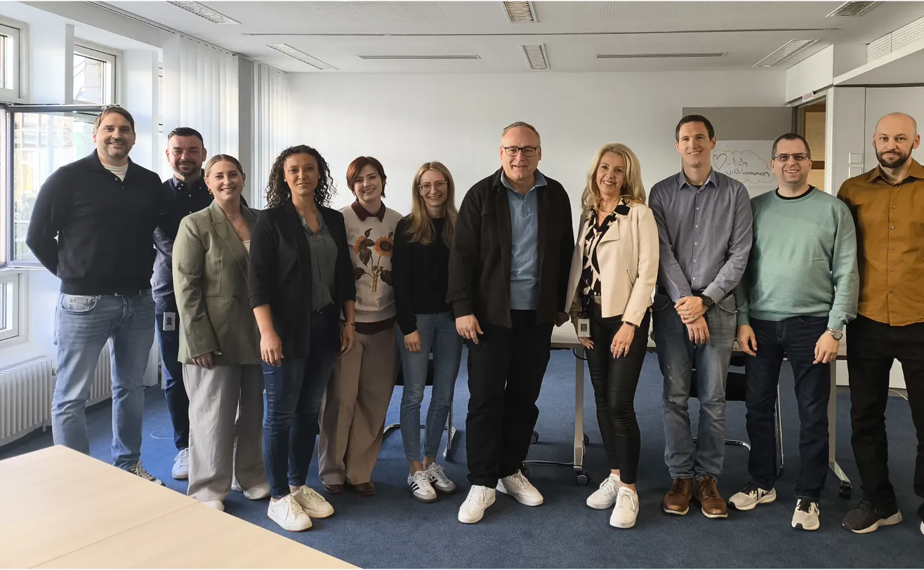 Gruppe von Männern und Frauen steht lächelnd nebeneinander in einem hellen Seminar- oder Besprechungsraum.