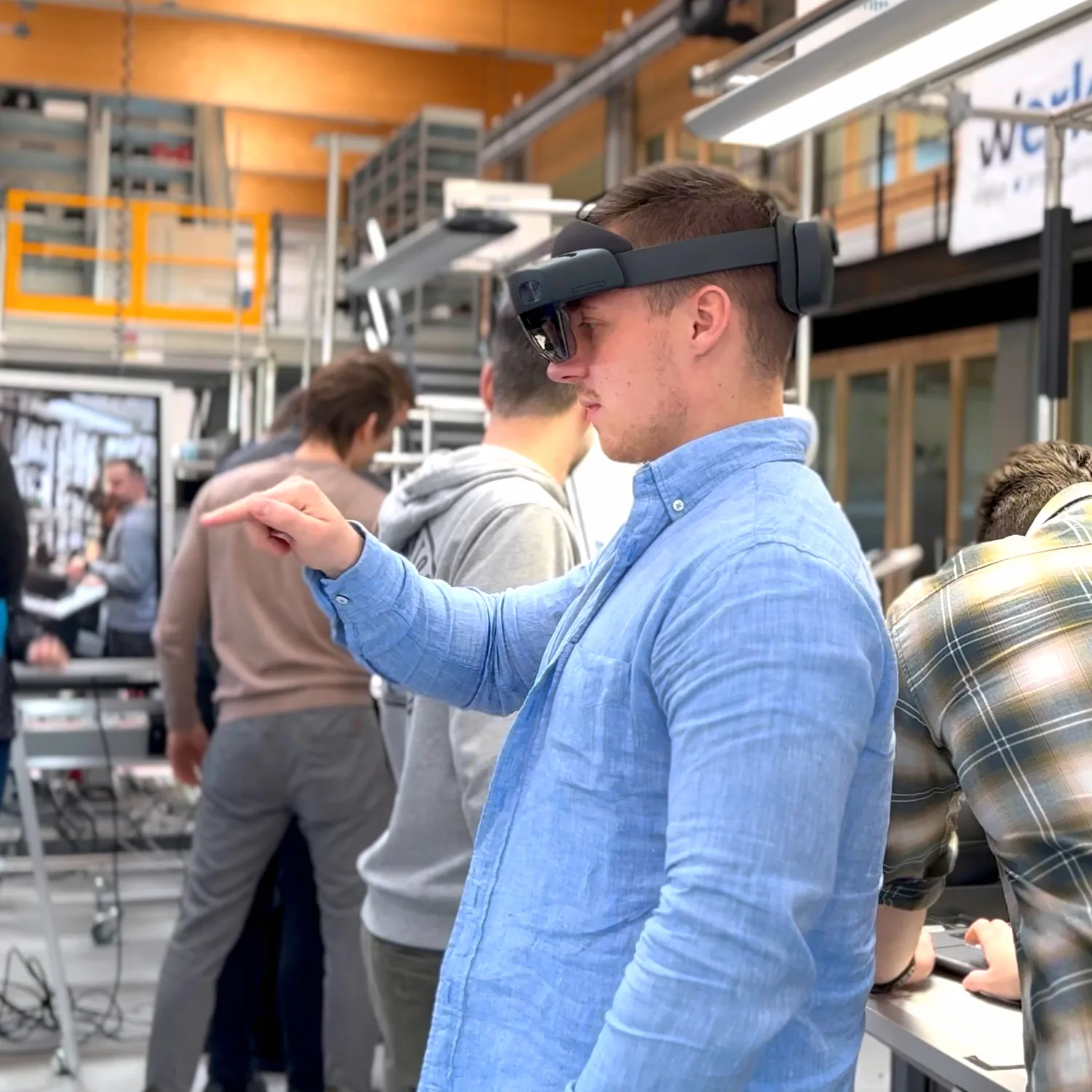 Junger Seminar Teilnehmer trägt AR-Brille und interagiert mit Gesten in einer technischen Werkstatt mit weiteren Personen.