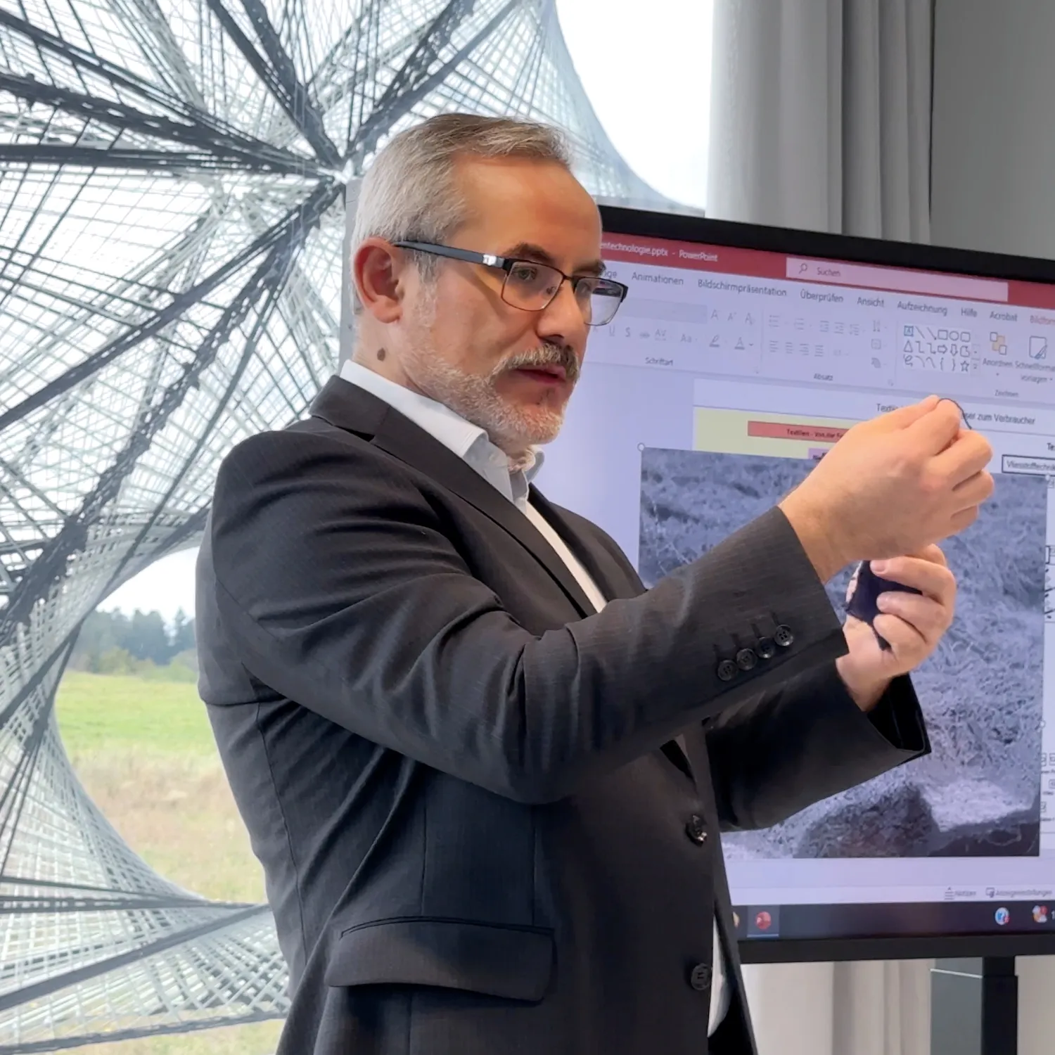 Älterer gut gekleideter Mann mit Brille und Anzug präsentiert Stoffsample vor einem Bildschirm mit PowerPoint-Folie.