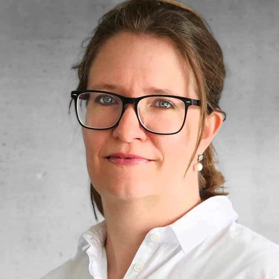 Portraitfoto von Verena Szembek, Frau mit Brille vor grauem Hintergrund.